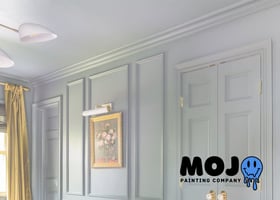 A light-blue room with matching walls and ceiling, featuring ornate wall paneling, a gold-framed floral painting under a picture light, and a modern white chandelier. Mojo Painting Company