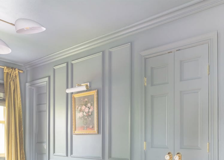 A light-blue room with matching walls and ceiling, featuring ornate wall paneling, a gold-framed floral painting under a picture light, and a modern white chandelier.