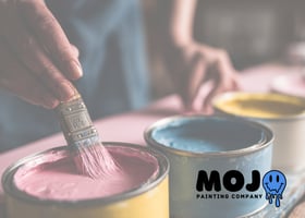 A hand dipping a paintbrush into a can of pink paint, with other open paint cans nearby and a small mojo painting company in the corner. phoenixville malvern
