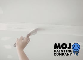 Hand using a putty knife to spackle and smooth a ceiling drywall seam, with the Mojo Painting Company logo in the corner.