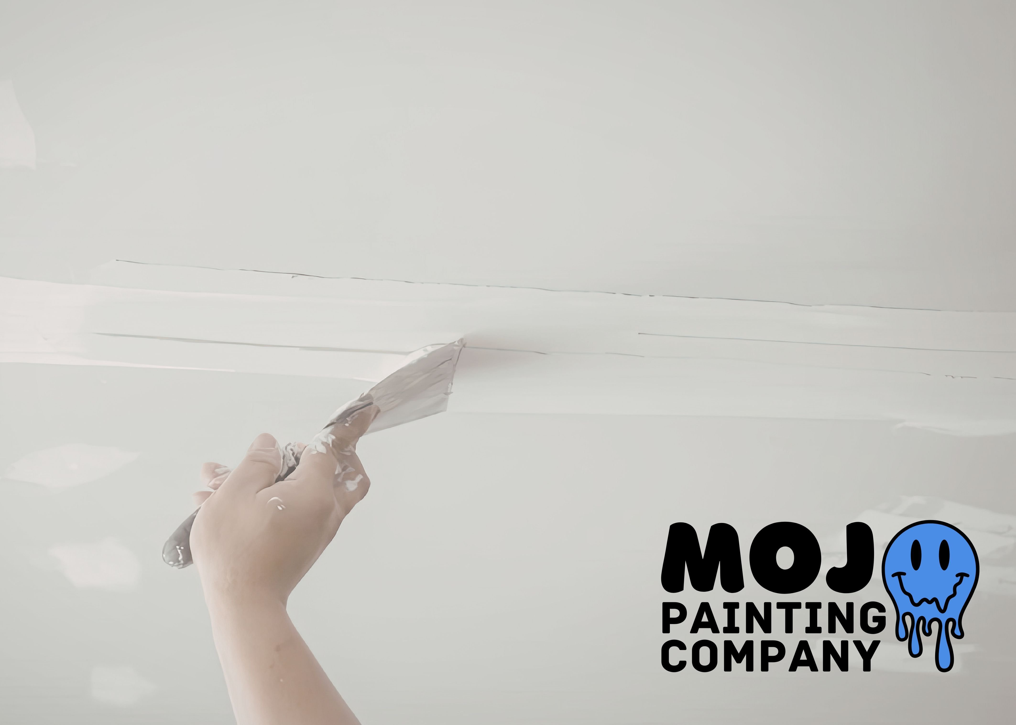 Hand using a putty knife to spackle and smooth a ceiling drywall seam, with the Mojo Painting Company logo in the corner.