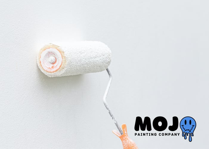 Paint roller applying fresh white paint to a smooth light gray wall, with the Mojo Painting Company logo in the bottom right corner featuring bold black text and a blue dripping smiley face icon.